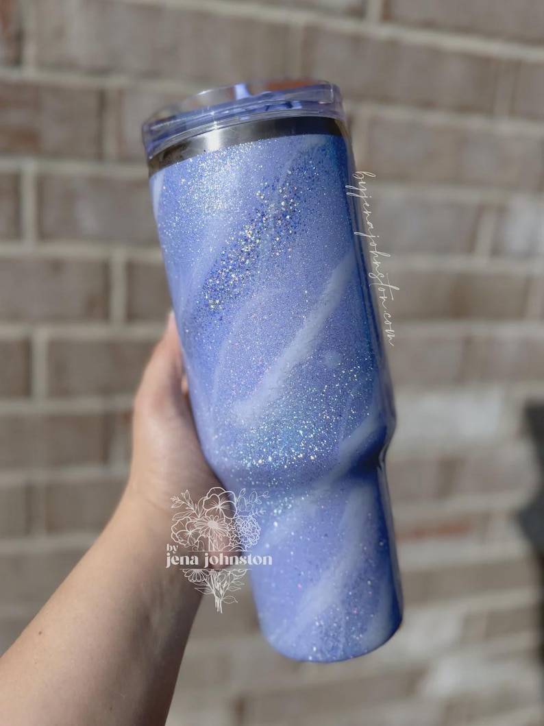 May include: A blue glitter tumbler with a swirling white design. The tumbler has a clear lid and is held by a hand. The text "by jenajohnston.com" is visible on the side. The tumbler is a stylish drinkware item.