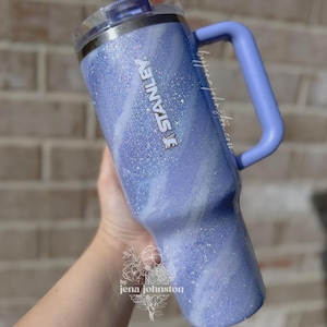 May include: A light blue Stanley tumbler with a handle and a clear lid. The tumbler has a glittery, marbled design with the Stanley logo in silver. The handle is a solid light blue. The tumbler is held by a person.