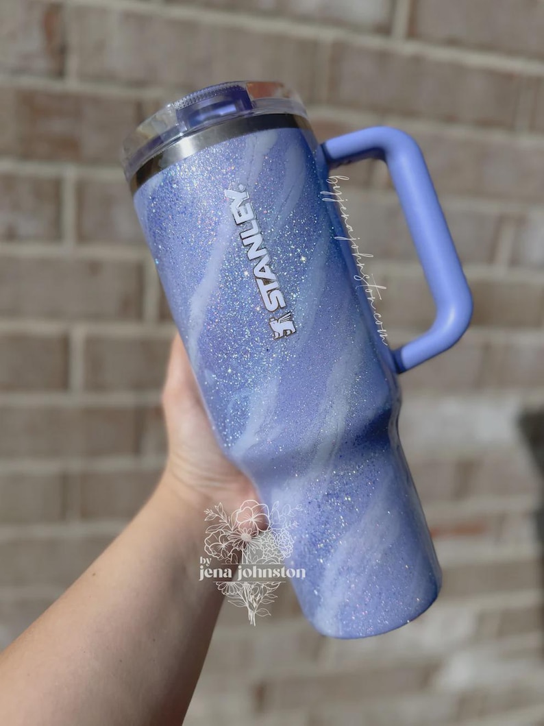 May include: A light blue Stanley tumbler with a handle and a clear lid. The tumbler has a glittery, marbled design with the Stanley logo in silver. The handle is a solid light blue. The tumbler is held by a person.