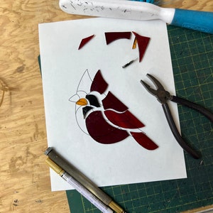 K&ouml;nnte beinhalten: Ein Buntglas-Kardinal in Rot-, Orange- und Schwarzt&ouml;nen wird auf einer gr&uuml;nen Schneidematte zusammengesetzt. Die Glasst&uuml;cke sind auf einem wei&szlig;en Blatt Papier angeordnet. Eine Zange, ein goldener Stift und ein Glasschneider sind ebenfalls sichtbar.