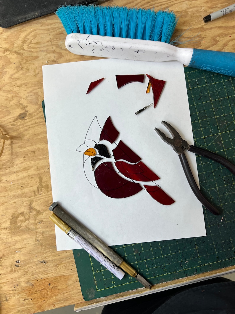 K&ouml;nnte beinhalten: Ein Buntglas-Kardinal in Rot-, Orange- und Schwarzt&ouml;nen wird auf einer gr&uuml;nen Schneidematte zusammengesetzt. Die Glasst&uuml;cke sind auf einem wei&szlig;en Blatt Papier angeordnet. Eine Zange, ein goldener Stift und ein Glasschneider sind ebenfalls sichtbar.