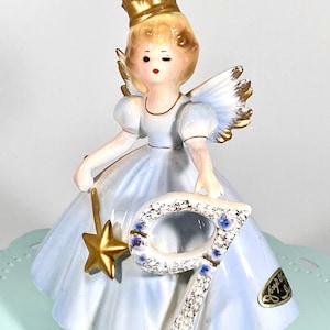 May include: A porcelain figurine of a girl wearing a blue dress and a gold crown, holding a gold star wand. She is standing next to a large, gold-trimmed number nine. The figurine is on a light blue background.