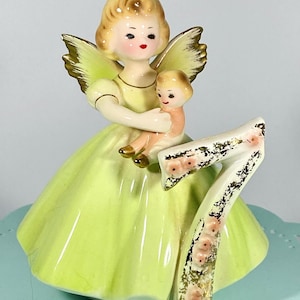 May include: A porcelain figurine of a girl angel with wings holding a doll. She is wearing a green dress and standing on a number seven with pink flowers. The number seven is gold with a glittery finish.