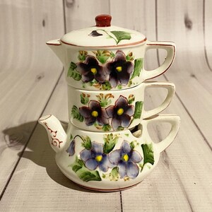 May include: A set of three white ceramic teacups with floral designs. The cups are stacked on top of each other, with the largest cup at the bottom and the smallest cup at the top. The cups are decorated with purple and green flowers.