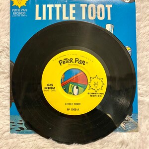 Op de afbeelding: Een zwarte vinylplaat met een geel label. Het label toont een cartoon trein met een glimlachend gezicht en de tekst "Little Toot PP 1009 A". De plaat maakt deel uit van de "Sunshine Series" en is een 45 RPM plaat.