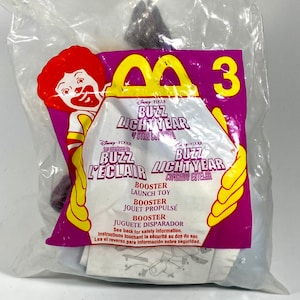 Op de afbeelding: Een paars Buzz Lightyear speelgoed uit de Disney Pixar film Toy Story. Het speelgoed is verpakt in een doorzichtige plastic zak met het McDonald's Happy Meal logo en de woorden "Buzz Lightyear" en "Booster Launch Toy".