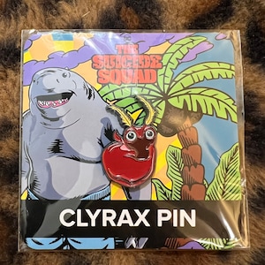 May include: A red and black enamel pin featuring the character Clyrax from the DC Comics The Suicide Squad movie. The pin is packaged on a card with a background image of the character King Shark and the text "The Suicide Squad". The card also includes the text "Clyrax Pin".