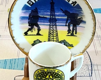 May include: A white ceramic teacup and saucer set with a gold rim. The saucer features a colorful image of an oil derrick and two Native American figures with the word "OKLAHOMA" in a gold circle. The teacup has a gold rim and a green and gold seal with the words "Great Seal of the State of Oklahoma" and "1907".