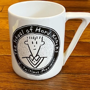 May include: White ceramic mug with a black and white illustration of a cartoon character and the text "The School of Hard Knocks - E. Twitimus. Maximus."