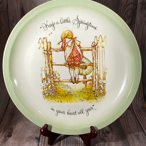 Könnte beinhalten: Ein grüner und weißer Keramikteller mit einem Mädchen in einem roten Kleid, das über einen weißen Lattenzaun schaut. Der Teller trägt den Text "Keep a little Springtime in your heart all year".