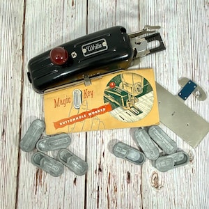 May include: An assortment of vintage sewing machine attachments and accessories. A black "White" brand buttonhole maker with a red knob, a "Magic Key Buttonhole Worker" box, and several silver-colored metal attachments are visible.