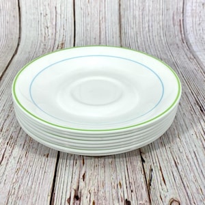 Puede incluir: Una pila de platillos de cer&aacute;mica blanca con un borde verde y azul. Los platillos est&aacute;n apilados sobre una superficie de madera.