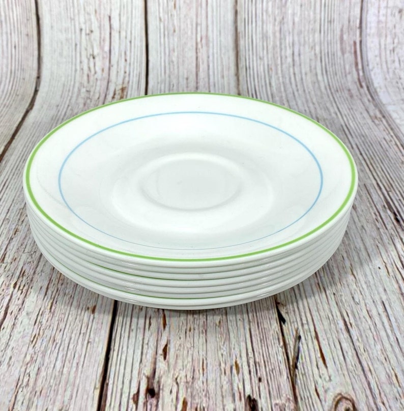 Puede incluir: Una pila de platillos de cer&aacute;mica blanca con un borde verde y azul. Los platillos est&aacute;n apilados sobre una superficie de madera.