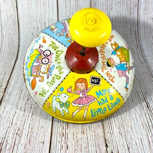 Könnte beinhalten: Vintage Kinderspielzeug mit gelbem Oberteil und roter Basis. Das kreisförmige Metallspielzeug zeigt Illustrationen von Kinderreimen, darunter "Mary Had a Little Lamb" und "Tom the Piper's Son."