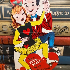 Può includere: Un biglietto di San Valentino vintage con una coppia di cartoni animati che pattina. Il ragazzo indossa una giacca rossa e un papillon blu, e la ragazza indossa una gonna gialla e una maglietta nera. Il biglietto dice "Fai un buon pattinaggio, sii il mio San Valentino!"