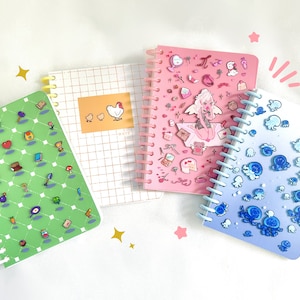Puede incluir: Cuatro cuadernos de espiral diferentes con varios diseños. Un cuaderno tiene una cubierta verde con un patrón de cuadrícula e imágenes pixeladas de artículos de videojuegos. Otro cuaderno tiene una cubierta rosa con un patrón de cuadrícula y una variedad de pegatinas. Un tercer cuaderno tiene una cubierta blanca con un patrón de cuadrícula y una ilustración de pollo. El cuarto cuaderno tiene una cubierta azul con un patrón degradado y pegatinas de pulpos azules.