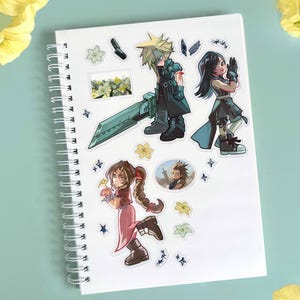 May include: White spiral-bound notebook adorned with colorful stickers featuring anime-style characters. The stickers depict figures with spiky blonde hair, black hair, and various outfits, along with floral and star-shaped accents. The notebook is on a light blue surface.