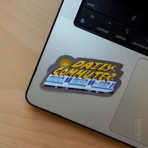 Puede incluir: Una pegatina gris y amarilla con el texto "Daily Commuter" y una imagen de dibujos animados de un tren. La pegatina est&aacute; en un port&aacute;til plateado.