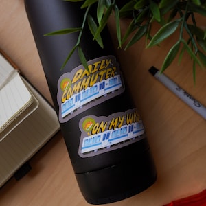 Puede incluir: Una botella de agua negra con dos pegatinas. La pegatina superior dice "Daily Commuter" con un tren de dibujos animados. La pegatina inferior dice "On My Way" con un tren de dibujos animados.