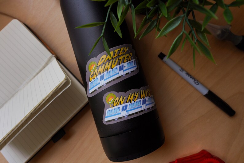Puede incluir: Una botella de agua negra con dos pegatinas. La pegatina superior dice "Daily Commuter" con un tren de dibujos animados. La pegatina inferior dice "On My Way" con un tren de dibujos animados.
