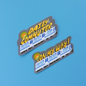 Puede incluir: Dos pegatinas con un tren de dibujos animados con el texto "Daily Commuter" y "On My Way!" en un estilo de c&oacute;mic colorido.