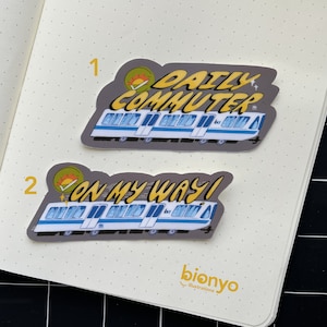 Puede incluir: Dos pegatinas con un tren de dibujos animados con el texto "Daily Commuter" y "On My Way!" Las pegatinas est&aacute;n en una p&aacute;gina de cuaderno blanca con puntos.