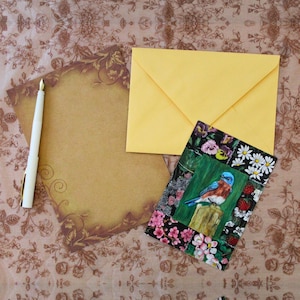 Op de afbeelding: Een witte vulpen, een vel beige briefpapier met bruine bloemenranden, een effen gele envelop en een rechthoekige kaart met een blauwe vogel op een paal, omringd door een collage van bloemen, bessen en bladeren.