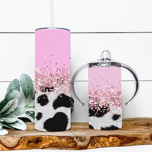 K&ouml;nnte beinhalten: Zwei rosa und wei&szlig;e Kuhmuster-Becher mit rosa Glitzerakzenten. Der gr&ouml;&szlig;ere Becher hat einen Strohhalm und der kleinere Becher hat Griffe.