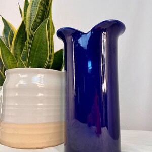 May include: A tall, cylindrical ceramic vase with a deep, glossy cobalt blue finish and a distinctively curved, undulating rim. The vase features a smooth, reflective surface that catches light, standing upright with a simple, clean, and modern structural form.