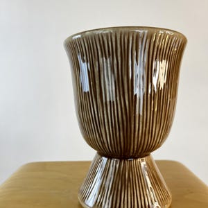 May include: A brown ceramic goblet with a glossy finish and a vertical ribbed texture. The vessel features a wide, rounded bowl that tapers down to a flared, pedestal-style base, creating a distinct silhouette with consistent vertical lines.
