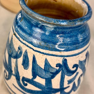 May include: A ceramic jar with a cream-colored base and hand-painted blue decorative patterns. The jar features a wide, rounded body, a thick rim, and a distinct blue calligraphic script encircling the exterior. The interior is glazed in brown.