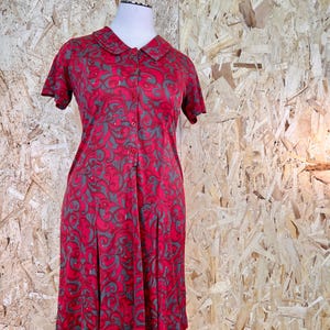 May include: A red dress with a grey swirling pattern is displayed on a mannequin. The dress has short sleeves, a collared neckline, and a button-down front. The skirt is pleated, and the dress appears to be made of a lightweight fabric.