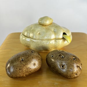 May include: A ceramic lidded container with a light yellow speckled finish and a small spoon, accompanied by two dark brown, oval-shaped ceramic salt and pepper shakers, each featuring small holes on the top surface for dispensing seasonings.