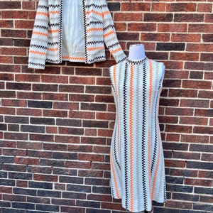 May include: A vintage two-piece outfit featuring a sleeveless dress and a matching jacket. The dress and jacket are white with vertical stripes in orange, black, and gray. The jacket is open-fronted with a zig-zag trim.