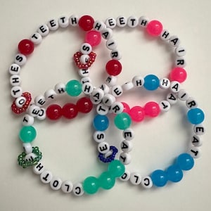 May include: Four beaded bracelets with white letter beads spelling out "TEETH," "HAIR," "CLOTHES," and "EAT." The bracelets feature red, pink, turquoise, and blue beads, with small decorative accents.