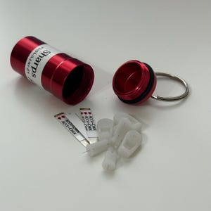 May include: A red metal sharps container with the word "Sharps" and "biohazard" printed on it. The container is open, revealing the inside. Several white and silver medical supplies and test strips are next to the container.