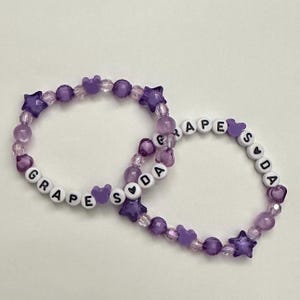 May include: Two beaded bracelets with the words "GRAPE SODA" spelled out in white letter beads. The bracelets feature purple beads, star-shaped beads, and heart-shaped beads. The bracelets are on a white background.