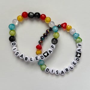 May include: Two beaded bracelets with the text "DISABLED AF". The bracelets feature a variety of colorful beads, including white letter beads, black beads, and heart-shaped beads. The beads are in shades of red, yellow, blue, green, and clear.