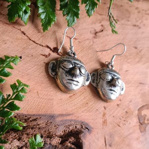 May include: A pair of silver-coloured earrings, each depicting a stylised face with notable features. The earrings hang from simple ear wires. The background is a wooden surface with green foliage.