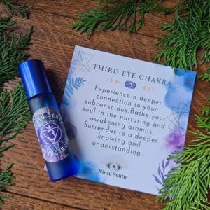 Könnte beinhalten: Eine blaue Rollerflasche mit einem Etikett mit einem lila Chakra-Symbol und dem Text "Nintu Sentu". Eine Karte mit den Worten "Third Eye Chakra" und Text über die Verbindung zu Ihrem Unterbewusstsein befindet sich neben der Flasche. Grünes Laub umgibt die Gegenstände.