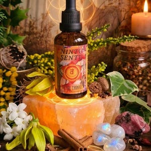 May include: A brown glass bottle with a black dropper, labeled "Nintu Sentu Sacral Chakra," sits atop an orange crystal. The image includes cinnamon sticks, white flowers, and a lit candle, with a glowing geometric design in the background.