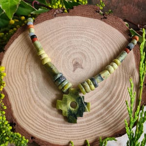 May include: A handmade necklace with a green stone pendant in a cross shape. The necklace has a beaded strand with green, yellow, black, red, and orange beads. The necklace is displayed on a wooden slice, with a natural background.