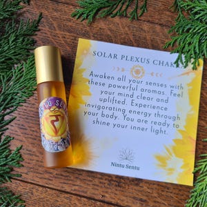 May include: A glass roller bottle with a gold cap and a label featuring a yellow chakra symbol and the text "Nintu Sentu". A card with text about the Solar Plexus Chakra is next to the bottle. The background includes greenery.