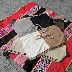 May include: A woven textile featuring a vibrant red, black, and pink geometric border pattern. Layered on top is a square fabric piece divided into four panels of black, grey, white, and tan, accompanied by a thin, multicoloured beaded cord.