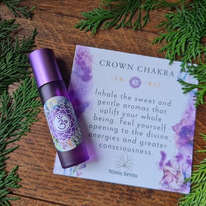 May include: A purple essential oil roller bottle with a label featuring a symbol and the words "Nintu Sentu" sits next to a card that reads "Crown Chakra". The card has text about inhaling aromas. Green foliage surrounds the items.