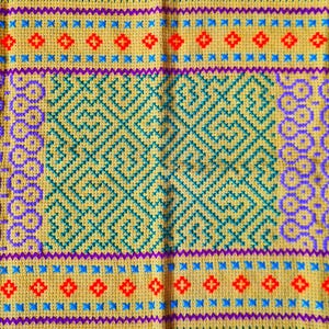May include: A rectangular, hand-stitched textile with a yellow background. The design features a central geometric pattern in teal, surrounded by borders of purple, red, and blue cross-stitch details. The edges are finished with a green border.