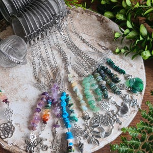 May include: A collection of silver tea infusers with decorative gemstone and charm accents. The infusers are arranged on a round, textured surface, surrounded by green foliage. The charms feature various shapes and colors.