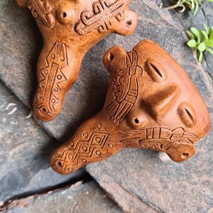 Może przedstawiać: Dwie brązowe ocariny ceramiczne z rzeźbionymi twarzami i geometrycznymi wzorami. Ocariny spoczywają na szarej kamiennej powierzchni, z zielonymi roślinami w tle. Unikalny, ręcznie robiony wygląd.