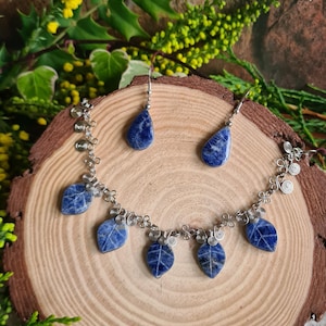 May include: A silver necklace and earring set with blue stone pendants and leaf-shaped charms. The jewellery is displayed on a wooden slice, surrounded by greenery and yellow flowers.