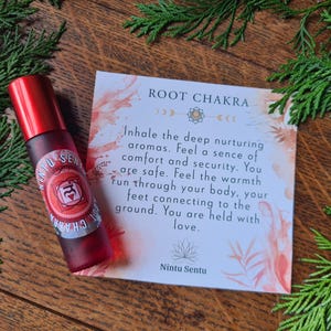 May include: A red essential oil roller bottle with a label that reads "Nintu Sentu Root Chakra" sits next to a card with text about the root chakra. The background includes green foliage on a wooden surface.